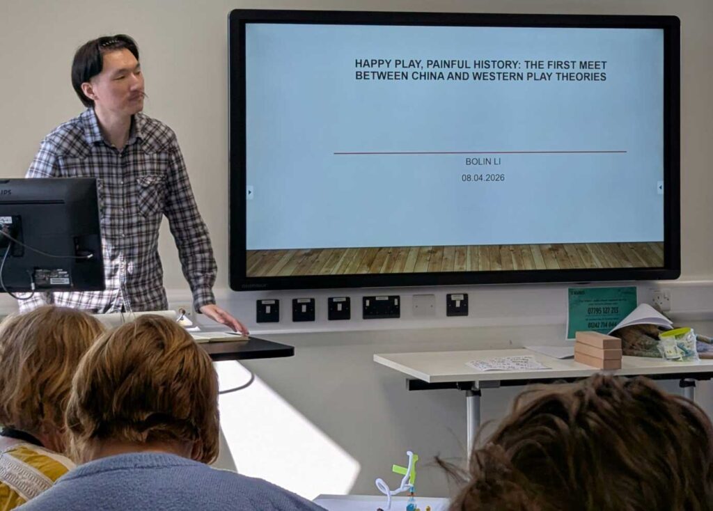 Bolin standing at the front of a seminar room giving a presentation entitled 'Happy Play, Painful History: the First Meet Between China and Western Play Theories'.