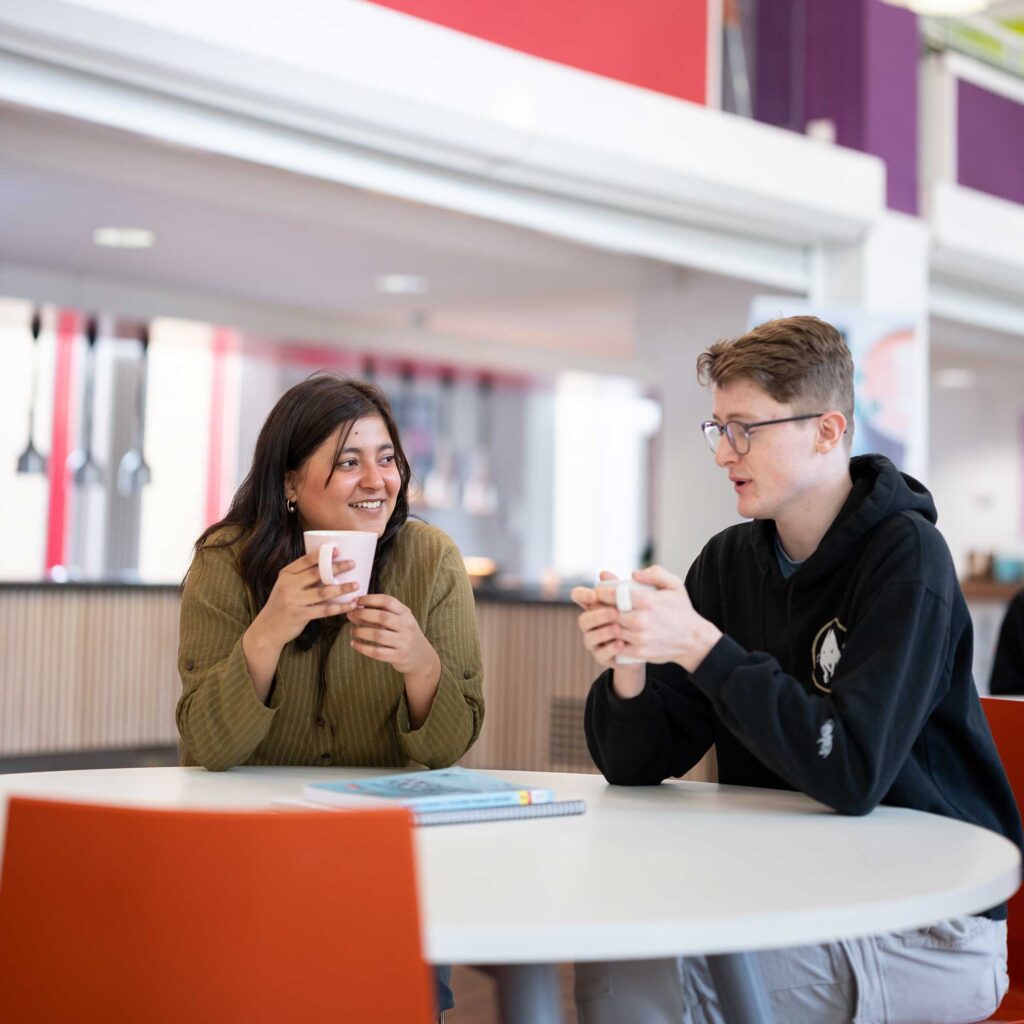 Two students sitting in the refectory at Francis Close Hall with mugs in their hands.