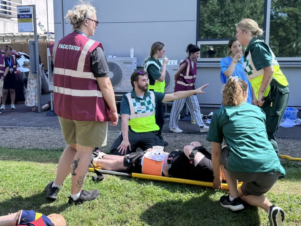 Paramedic Science students and facilitators take part in an outdoor major incident simulation, treating a person lying on a stretcher while others give directions and observe.