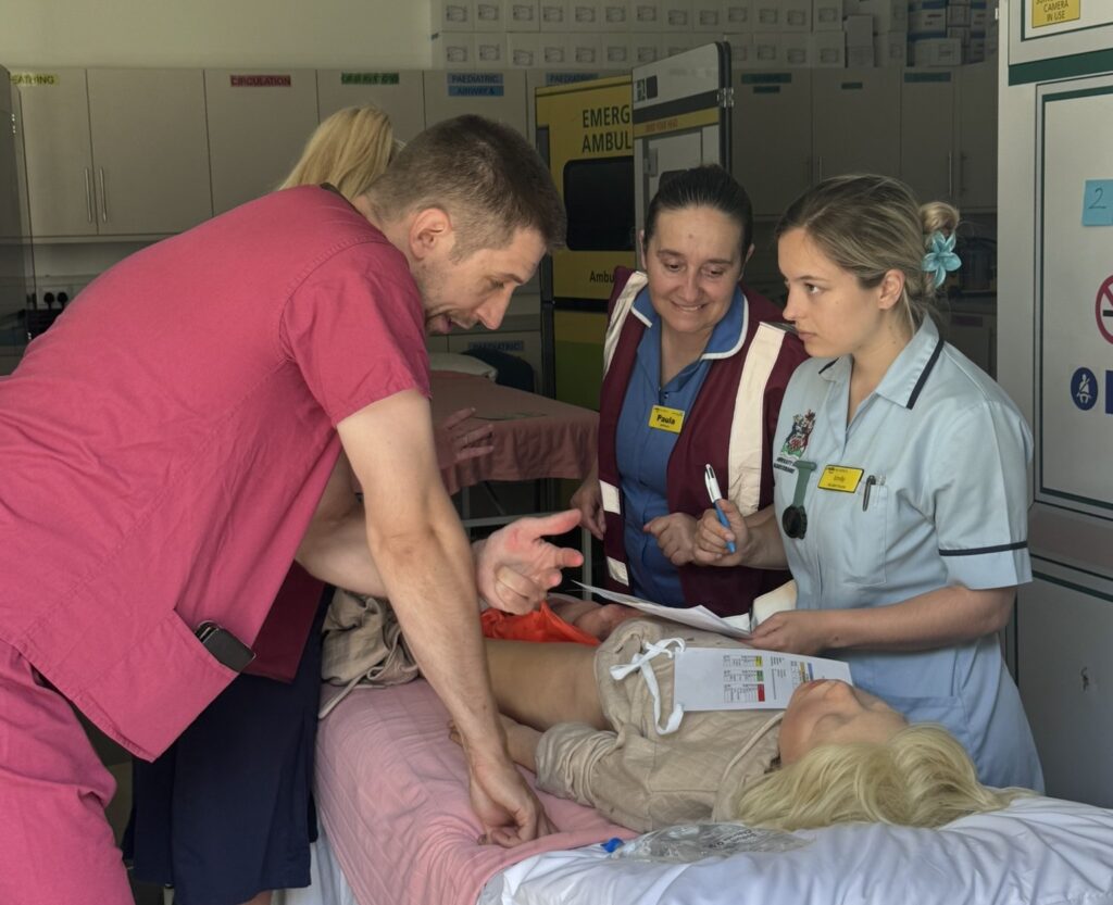 A healthcare simulation scene showing staff and a nursing student assessing a patient on a hospital bed, with one clinician explaining treatment while another records notes.