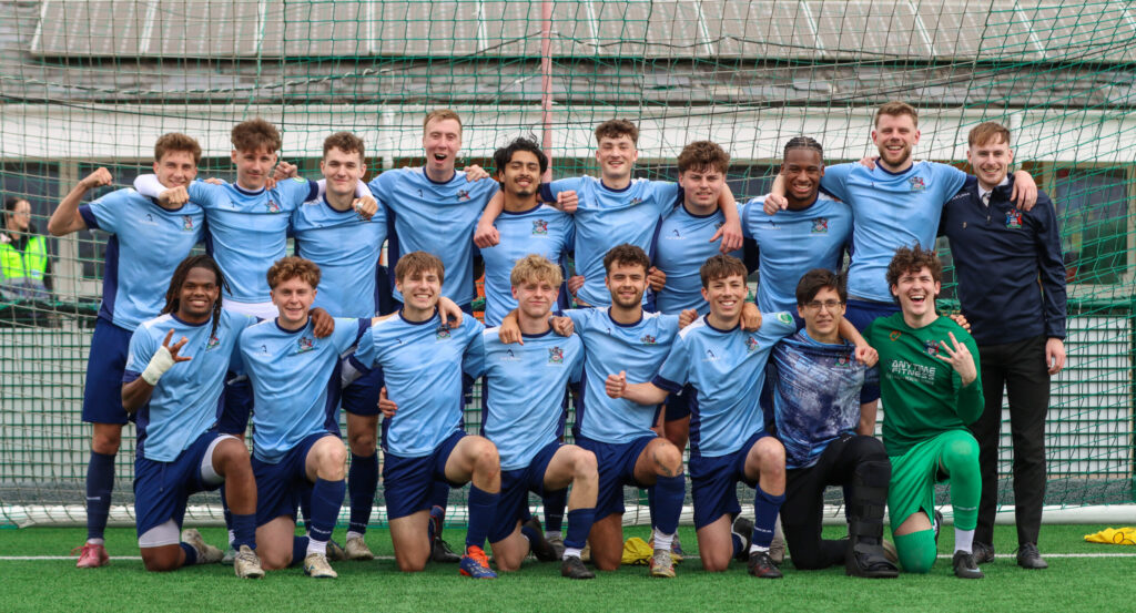 A men’s football team poses for a celebratory group photo on an outdoor pitch in front of a goal net, wearing mostly light blue jerseys and navy shorts, with one goalkeeper in green and a coach standing at the side.