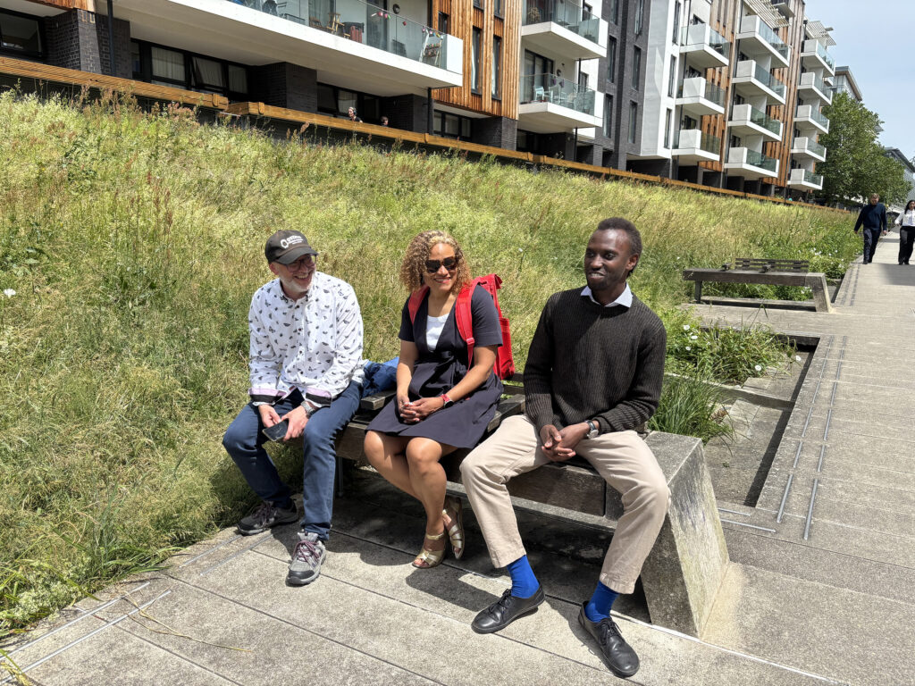 Three people sat on a bench with a grass verge and flats in the background