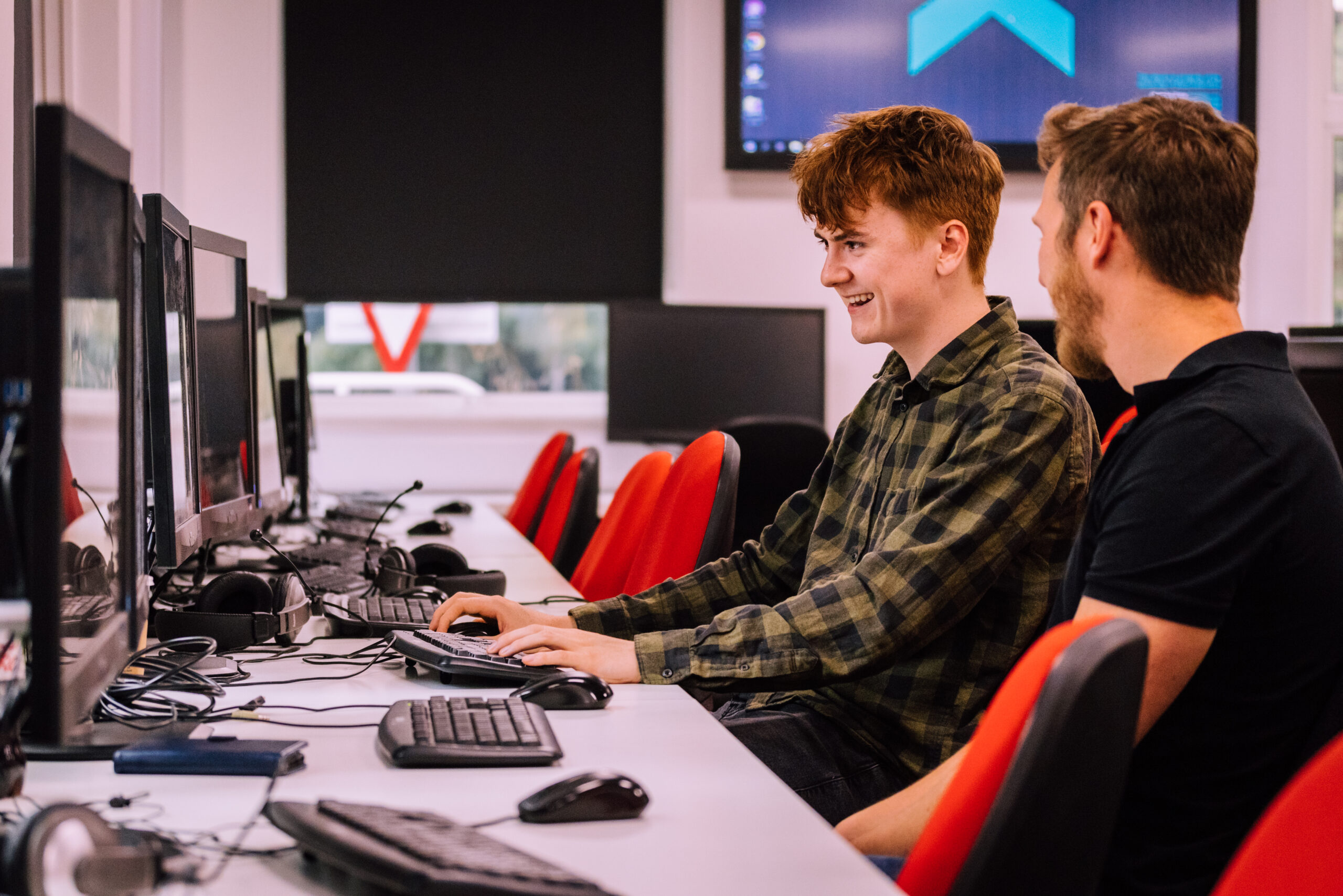 Two people sit at desktop computers in a university computer lab. A student in a green checked shirt smiles while using a keyboard, as a staff member beside him offers guidance. Multiple monitors, keyboards, headsets, and red-and-black chairs are visible.