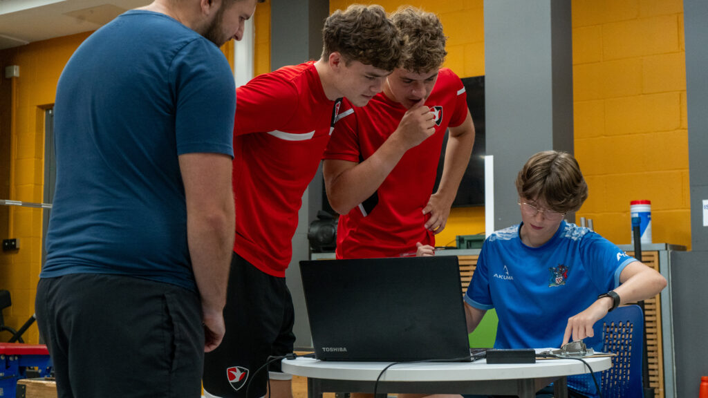 Four people gather around a laptop and electronic equipment in a sports science lab. Two Cheltenham Town players in red sports shirts lean in to observe while a seated student in a blue shirt works with the equipment, supervised by an adult in a blue T-shirt.