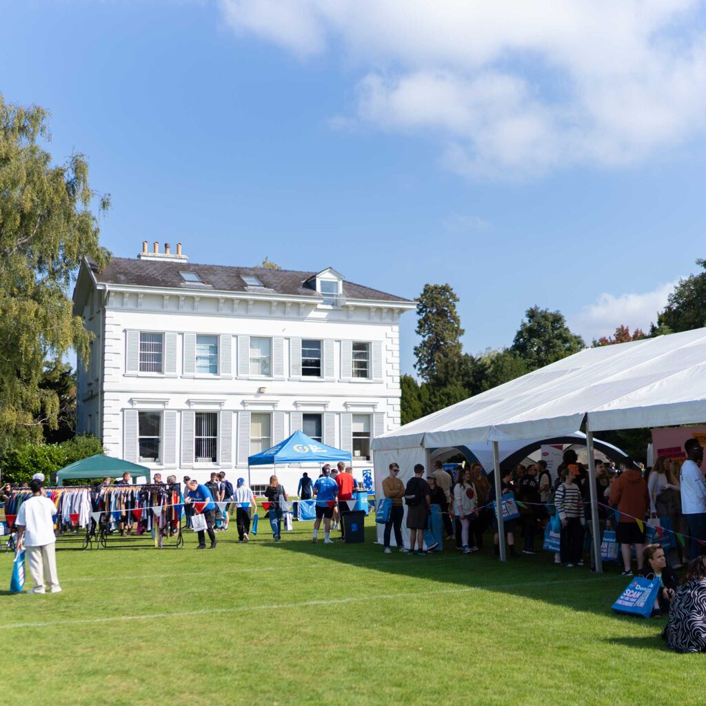 A view of the Welcome Fayre at Park campus showing a campus building and a large marquis on the grass and students looking at stalls.