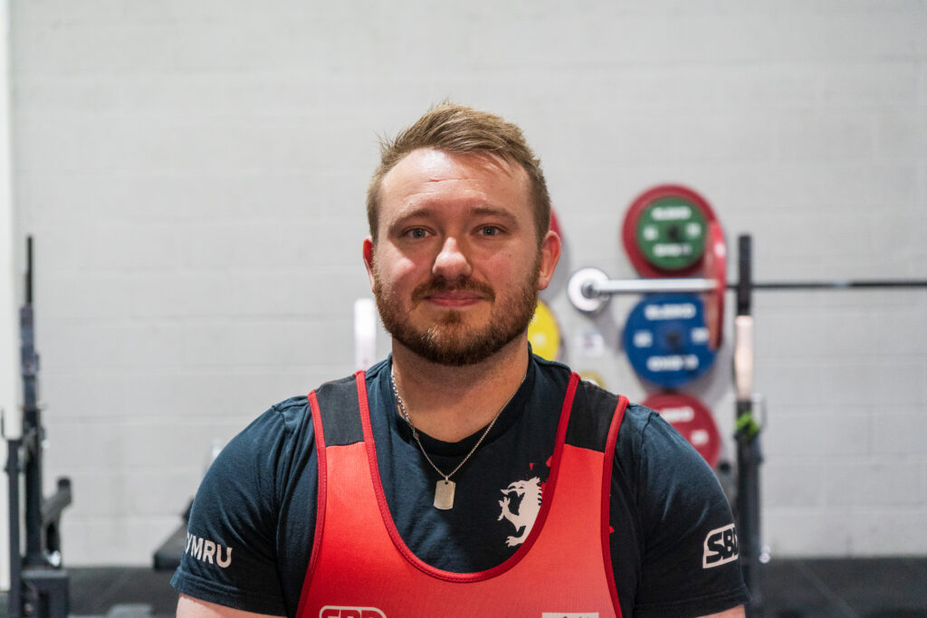 A male powerlifter wearing a red lifting singlet looks at the camera in a gym, with a barbell and coloured weight plates behind him.