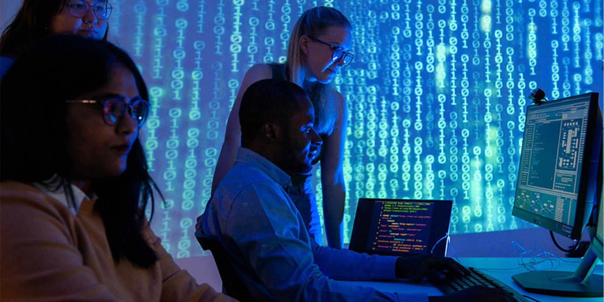 Four people work at computer stations in a dimly lit office, with blue binary code projected across the walls and screens displaying software and coding interfaces.