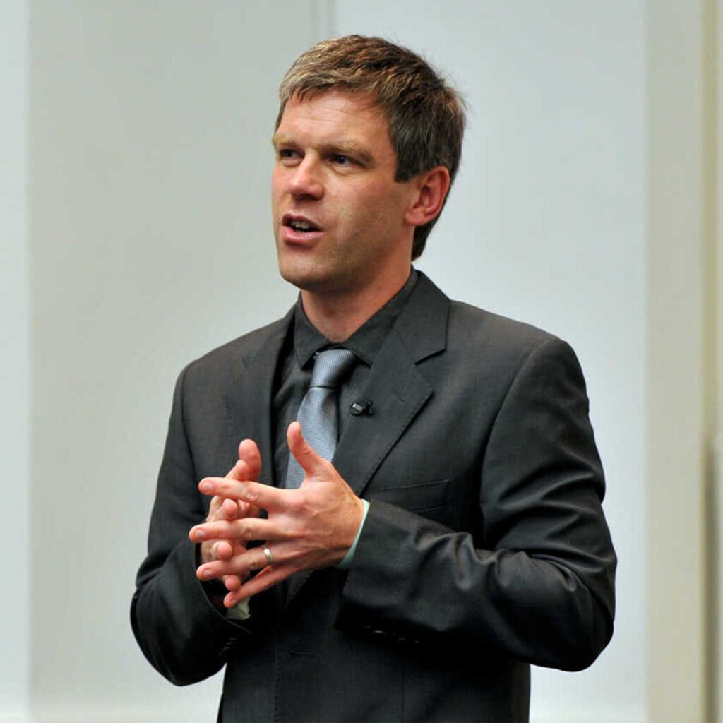 Damian Maye wearing a grey suit and tie addressing an audience with his hands clasped.