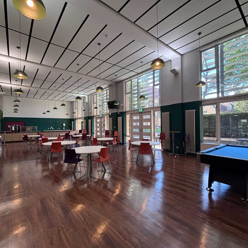 A photo on the food and social space at the refectory at FCH campus. Round tables and chairs in the centre of the room, a pool table in the foreground and food serving area in the background.