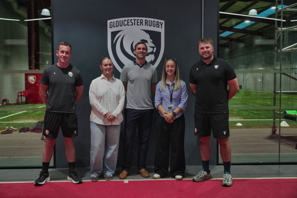 Five people stand together in front of a large Gloucester Rugby logo at an indoor rugby training facility. Two men wear Gloucester Rugby training kits, while the other three are dressed in smart-casual clothing.