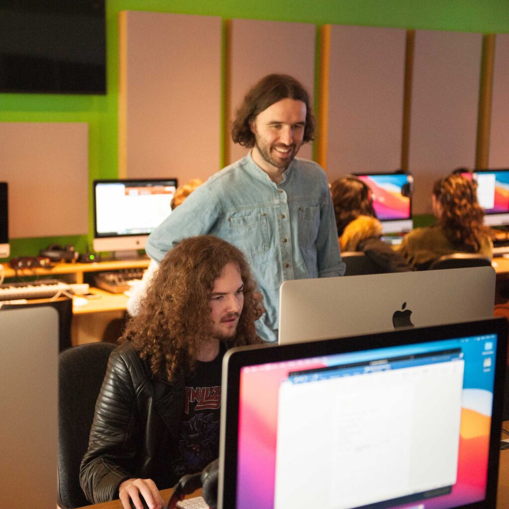 A lecturer standing behind a student who's sitting down and working at a computer.