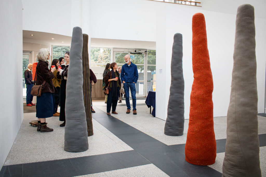Visitors chatting at an art exhibition in a bright, modern gallery space, surrounded by tall, tapered textile sculptures in shades of grey, brown and vibrant orange. The sculptures stand upright at varying heights, while groups of people gather and converse in the background.