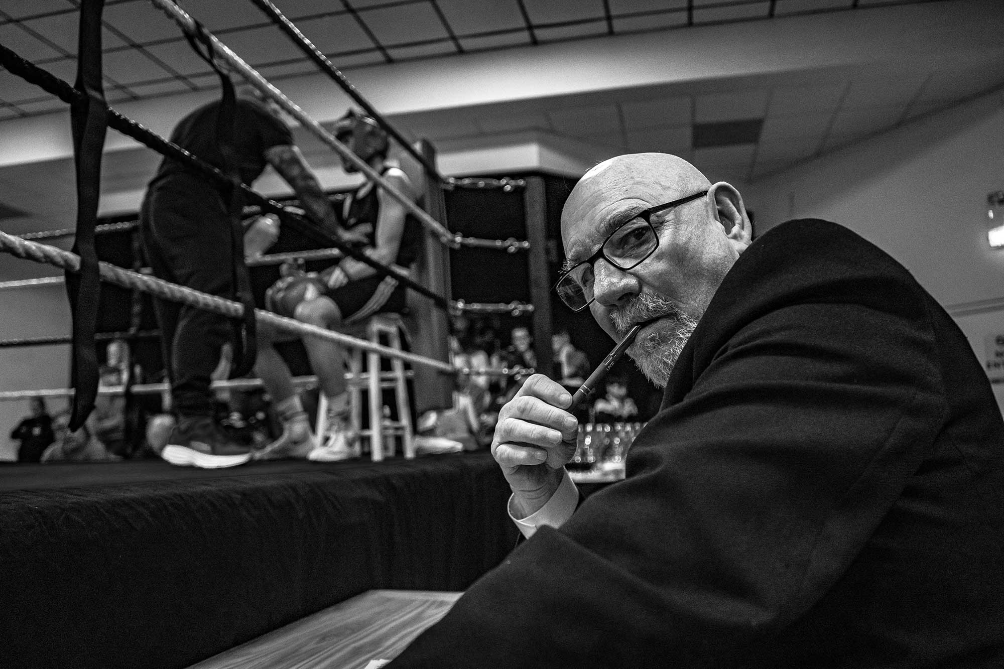 A man wearing a suit and smoking a pipe looks at the camera sternly while a boxing match takes place.