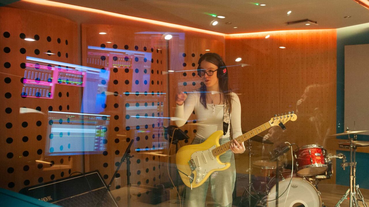 Music student with a guitar stands behind glass in a recording studio.